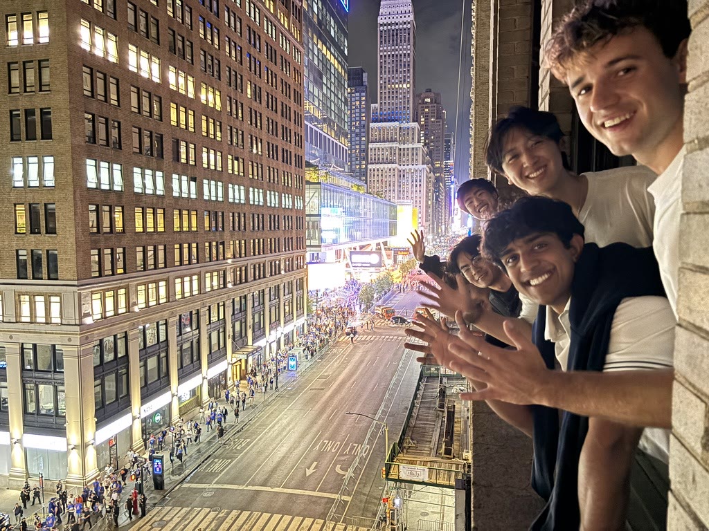 Friends leaning from a window above Seventh Avenue with the Empire State Building behind them