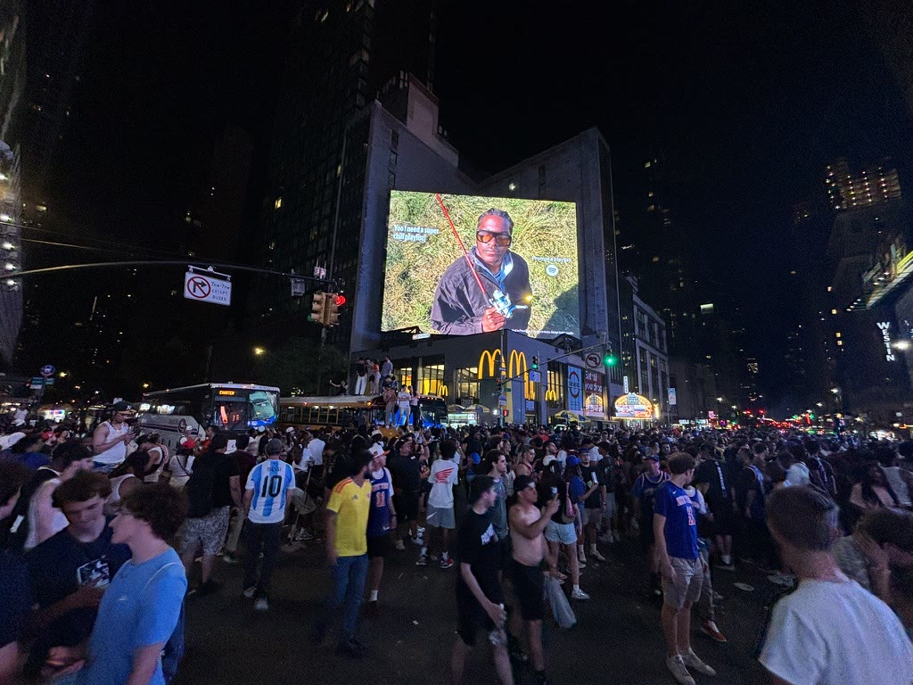 Knicks fans celebrating beneath a large screen on a Midtown street